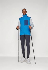 Mėlynas fliso liemenė su pilkais rankovėmis, krūtinės kišenė su užtrauktuku ir dvi žygeivių lazdos. Dėvima su tamsiomis tamprėmis ir baltais sportbačiais.