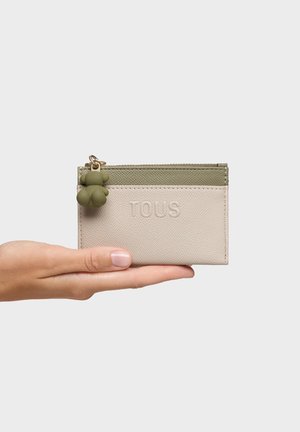 Mano sosteniendo un pequeño tarjetero TOUS beige y verde oliva con cremallera y un charm en forma de oso verde.