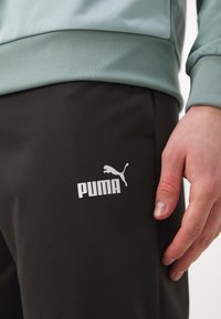 Sorte træningsbukser med et hvidt Puma-logo på låret, der har en glat tekstur. En lys blå t-shirt er lagd ovenpå bukserne.