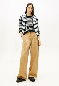 Gestreiftes Langarmoberteil mit blauen Akzenten, kombiniert mit weit geschnittenen, beigen Hosen und schwarzen klobigen Schuhen; das Model hat glattes braunes Haar.