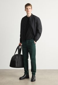 Chaqueta bomber negra sobre una camiseta negra, pantalones verdes y botas negras. Lleva un bolso de lona negro. Diseño simple y moderno.