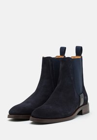 Paire de bottines Chelsea bleu marine en daim avec panneaux latéraux élastiques, languettes de tirage et petits talons en bois marron clair sur fond clair.