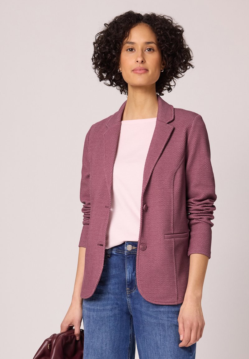Femme aux cheveux bouclés portant un blazer texturé mauve, un haut rose clair, un jean bleu, tenant un sac en cuir bordeaux plié devant un fond beige.