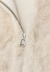 Beige konstfuskpälsjacka med silverdragkedjedetalj, som har en mjuk textur och ett slätt tygfoder synligt nära den öppna dragkedjan.