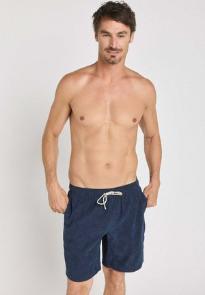 Homme torse nu aux cheveux courts et foncés portant un short bleu marine à cordon, debout les mains dans les poches devant un fond clair uni.