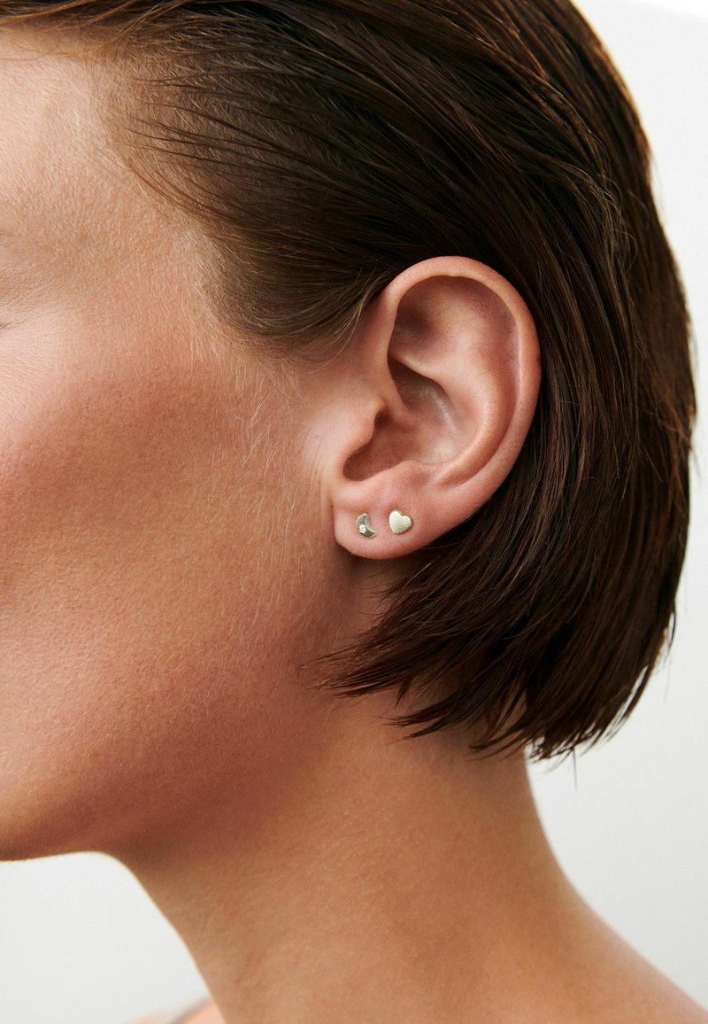 Gros plan sur l'oreille d'une femme aux cheveux courts et bruns, portant de petites boucles d'oreilles clous argentées en forme de croissant de lune et de cœur.