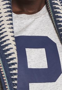 Grå tröja med en stor marinblå "P"-grafik. Bakgrunden har en stickad mönstrad sjal i marin, krämfärg och pastellfärger.