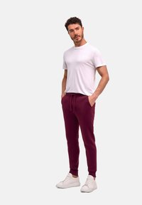 Burgunderfarbene Sweatpants mit Kordelzugbund, gerippten Bündchen, kombiniert mit einem schlichten weißen T-Shirt und weißen Sneakers. Glatter Stoff, lockere Passform.