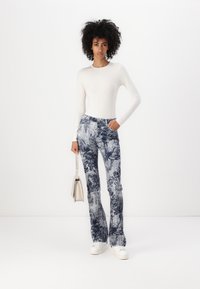 Flare jeans i blå og hvit blomstermønster med høy midje, kombinert med en hvit langermet topp og hvite sneakers, mens hun holder en sølvveske.