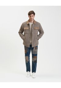Giacca a motivi a quadri marroni con due tasche sul petto, abbinata a jeans blu con vari motivi di toppe in marrone e nero. Sneakers bianche.