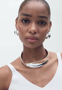 Collana a choker argentata con un design liscio e curvo, abbinata a orecchini a cerchio lucidi. La modella indossa un top bianco.