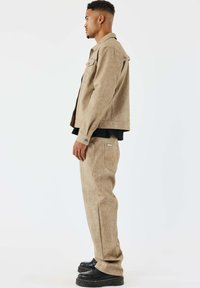Chaqueta marrón texturizada y pantalones a juego, ambos con un corte relajado. Los pantalones tienen un bolsillo con un sutil logotipo. Zapatos de plataforma negros.