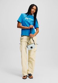 Plavi skraćeni majica s bijelim natpisom, uparena s blagim žutim cargo hlačama. Crna i krem crossbody torba s kvadratnim logom i podesivim remenom.