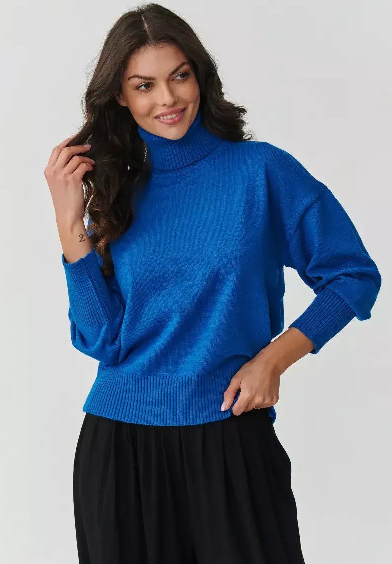 Niebieski sweter z dzianiny z wysokim kołnierzem i ściągaczami. Luźny krój i miękka tekstura. Noszony z czarnymi spodniami.