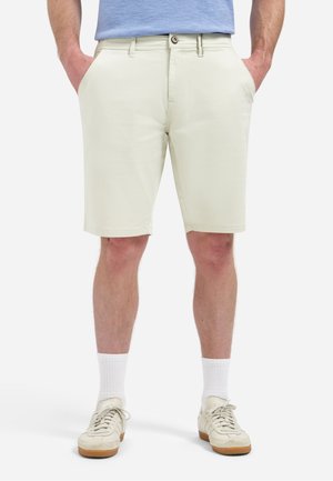 Man draagt licht beige knielange shorts, witte sokken en crèmekleurige sneakers, met handen in zakken tegen een effen achtergrond.