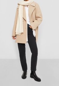 Abrigo de lana beige con solapas oversize, camisa blanca, pantalones negro y botas negras de tobillo. Acentuado con una bufanda de patrón ligero.