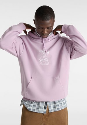 Sudadera rosa claro con bolsillo canguro y un diseño gráfico blanco en el pecho. Llevada sobre una camisa de cuadros azul y blanca y pantalones marrones.
