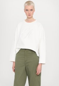 Sweatshirt blanc avec un logo "EH" en relief subtil, à la coupe ample et aux épaules tombantes, associé à un pantalon cargo vert olive.