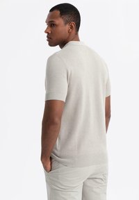 Camisa polo de punto de manga corta en beige claro con un patrón texturizado. Puños y dobladillo acanalados, llevada con pantalones de color claro. Vista posterior.