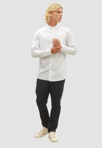 Weißes Hemd mit hohem Kragen und langen Ärmeln; kombiniert mit dunklen Hosen und hellen Turnschuhen; glattes Material und lässiges Design.