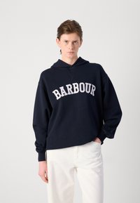 Sweatshirt navy com capuz, com "BARBOUR" em letras brancas atravessando o peito. Corte descontraído, mangas longas e tecido macio. Combinado com calças brancas.