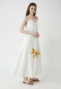 Weißes Maxikleid mit gelben Spaghettiträgern, das einen fransigen Ausschnitt und ein besticktes gelbes Palmenblattdesign nahe dem Saum hat.