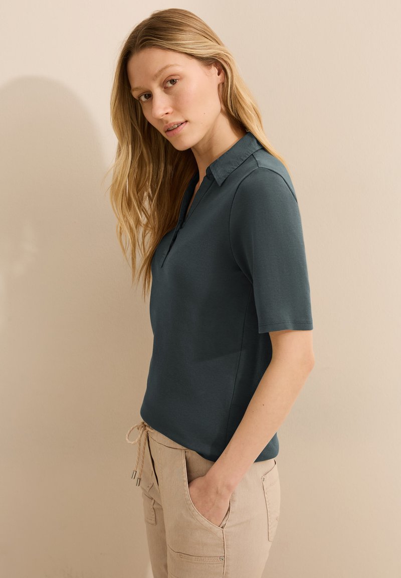 Tyrkysové polo tričko s límečkem a krátkými rukávy, vyrobené z měkké tkaniny. Spárováno s béžovými kalhotami s viditelnými kapsami.