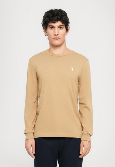 Un jeune homme avec des cheveux bouclés et foncés portant une chemise à manches longues beige avec un petit logo de cheval blanc sur la poitrine et un pantalon foncé.