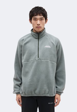 Homme portant un pull polaire gris à demi-fermeture éclair et un pantalon noir, debout devant un fond clair uni, faisant face à l'avant.