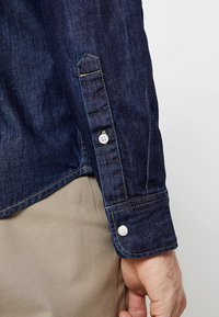 Jeansowa koszula z ciemnoniebieskiego materiału, zapinane mankiety, kontrastowe żółte przeszycia oraz kieszeń z klapką na rękawie.