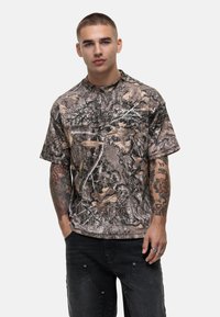Camo-patterned short-sleeve shirt in earthy tones with leafy designs. Features a round neck and relaxed fit. Accented logo on the chest.