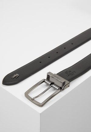 Zwart geperforeerde leren riem met zilverkleurige metalen gesp, met het Lacoste-logo in reliëf en een klein krokodillenembleem nabij het puntige uiteinde.