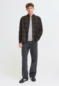 Camisa de franela a cuadros en marrón oscuro y negro, abotonada, con una camiseta gris debajo; combinada con unos vaqueros de mezclilla negros y zapatillas negras.