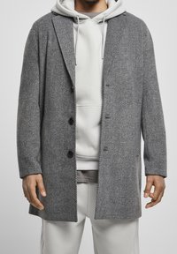 Grå hørindstrikjakke med klapkrave, enkelt knaplukning foran og tre knapper. Båret over en lysegrå hættetrøje og sweatpants.