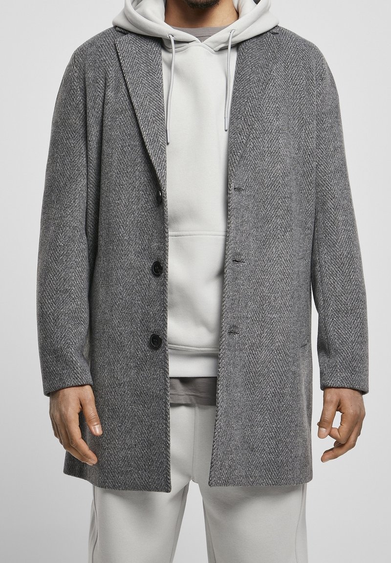 Grå hørindstrikjakke med klapkrave, enkelt knaplukning foran og tre knapper. Båret over en lysegrå hættetrøje og sweatpants.