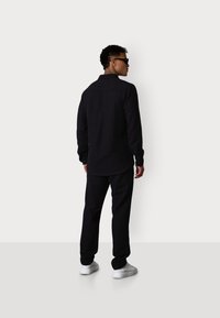 Schwarzes langärmliges Hemd mit Knopfmanschetten, kombiniert mit schwarzen Hosen und weißen Sneakers. Model steht mit dem Rücken zur Kamera und zeigt ein einfaches, elegantes Design.