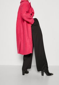 Cappotto in misto lana fucsia con ampio colletto e un solo bottone, abbinato a pantaloni neri a gamba larga e stivaletti con tacco. Tessuto strutturato.