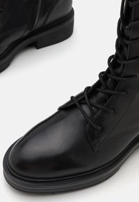Bottines en cuir noir à lacets, avec un bout rond, une surface texturée et une semelle plate en caoutchouc. Elles présentent des lacets robustes et des coutures renforcées.