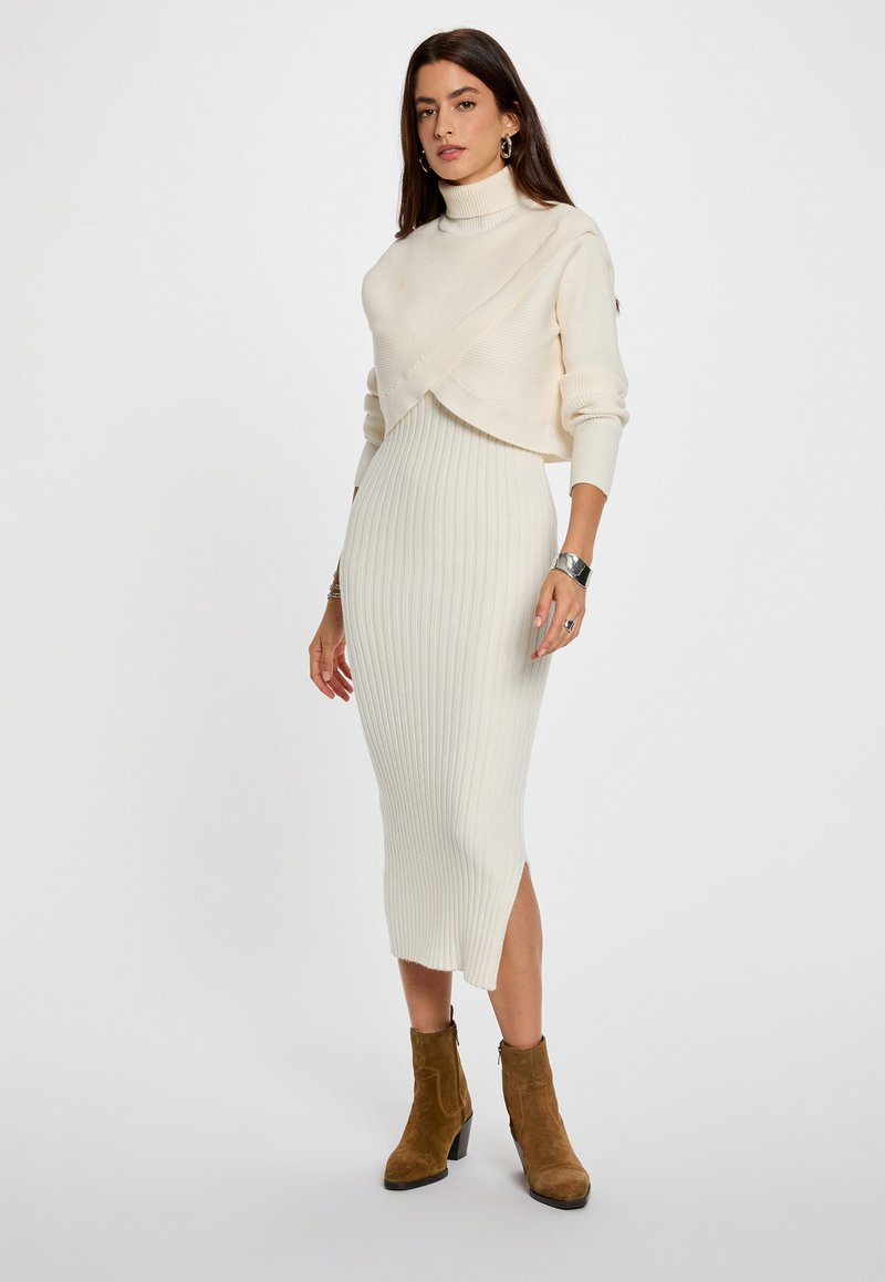 Abito in maglia a coste avorio con una silhouette aderente e uno spacco laterale, abbinato a un maglione a collo alto corto dello stesso colore. Stivali in suede marrone alla caviglia.
