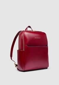 Rode leren rugzak met een gladde afwerking, voorzien van een voorvak met ritssluiting en een verstelbare riem. Gouden hardware accenten zijn zichtbaar.