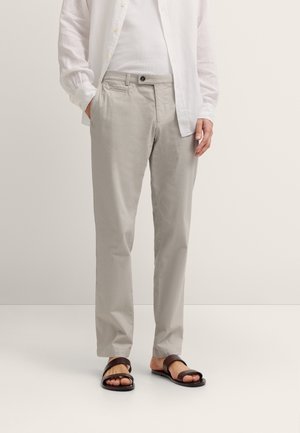 Man wearing light gray trousers, white shirt, and dark brown sandals standing against a plain light background.