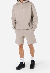 Sweat à capuche beige avec demi-fermeture éclair, poche avant, assorti à un short assorti. Porté avec des baskets noires et des chaussettes montantes blanches de marque.