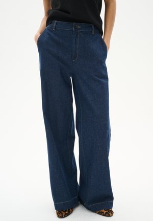 Femme portant un jean bleu foncé à jambes larges, les mains dans les poches, et des chaussures pointues à imprimé léopard visibles sous l'ourlet.