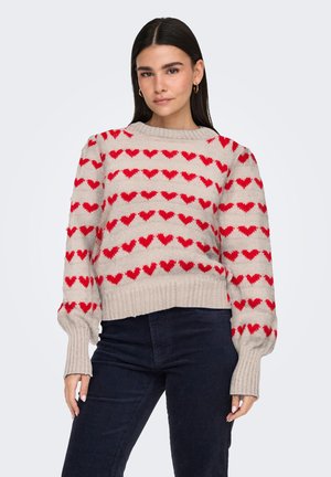 Femme aux longs cheveux foncés, portant un pull beige avec des motifs de cœurs rouges et un pantalon foncé, debout devant un fond clair uni.