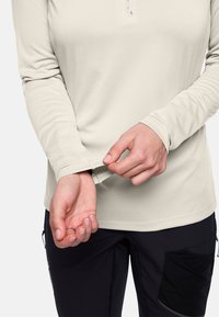 Persoon die de mouw van een beige lange mouwtop aanpast, gekleed in zwarte broek met zijzak, tegen een effen achtergrond.