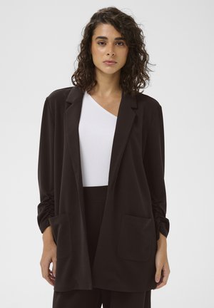 Femme aux cheveux bouclés porte un blazer noir ouvert avec deux poches avant par-dessus un top blanc à une seule épaule sur un fond clair uni.