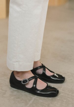 Chaussures Mary Jane en cuir verni noir avec design à bout fendu et boucle ovale décorative, portées avec un pantalon blanc cassé sur un sol beige.