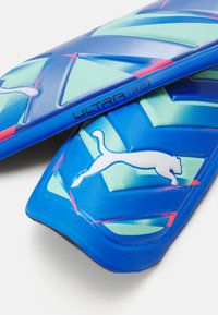 Plave zaštitne štitnike za potkoljenice s grafičkim dizajnom koji sadrži zelene naglaske i bijeli logotip pantere. Tekst glasi "ULTRA TWIST." Tekstura površine.