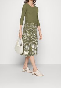 Abito a maglia a coste verde oliva con motivo astratto bianco, lunghezza fino al polpaccio, abbinato a mule con tacco color crema e una borsa a spalla.