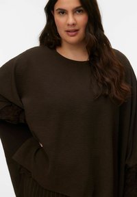 Haut marron texturé avec un col rond et une coupe oversize. Présente des manches superposées et un détail de poche latérale, confectionné dans un tissu doux.